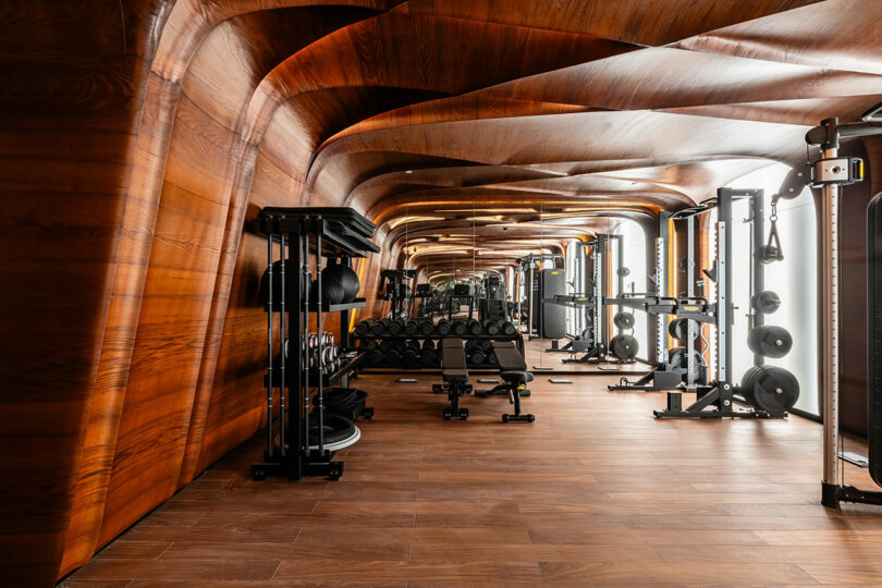 Modern gym interior with wooden ceiling and flooring, featuring various exercise equipment like weights and machines, lined up symmetrically along the walls.