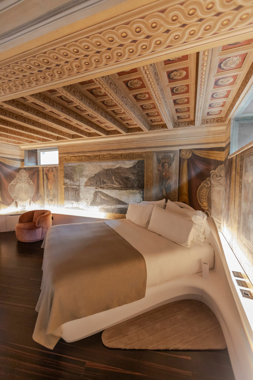 Luxurious hotel room with ornate ceiling and wall frescoes, featuring a modern bed, neutral bedding, and a plush chair on dark wood flooring.