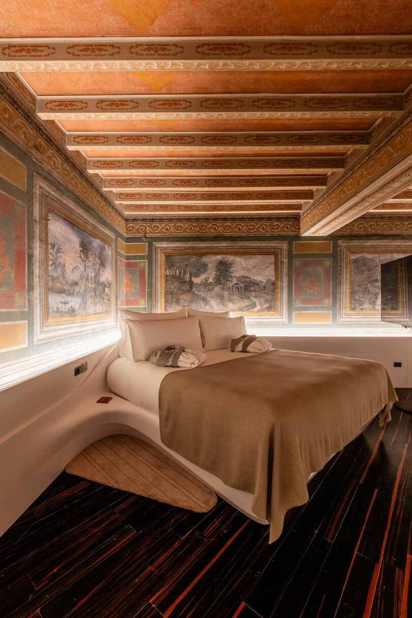 A bedroom with a large bed, beige bedding, and elaborate wall and ceiling paintings. The room has warm lighting and a dark wood floor.