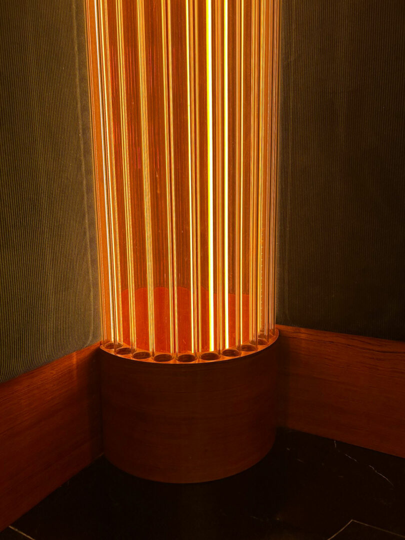 A warm, glowing cylindrical lamp is set in the corner between two textured walls, casting vertical lines of light and shadow.