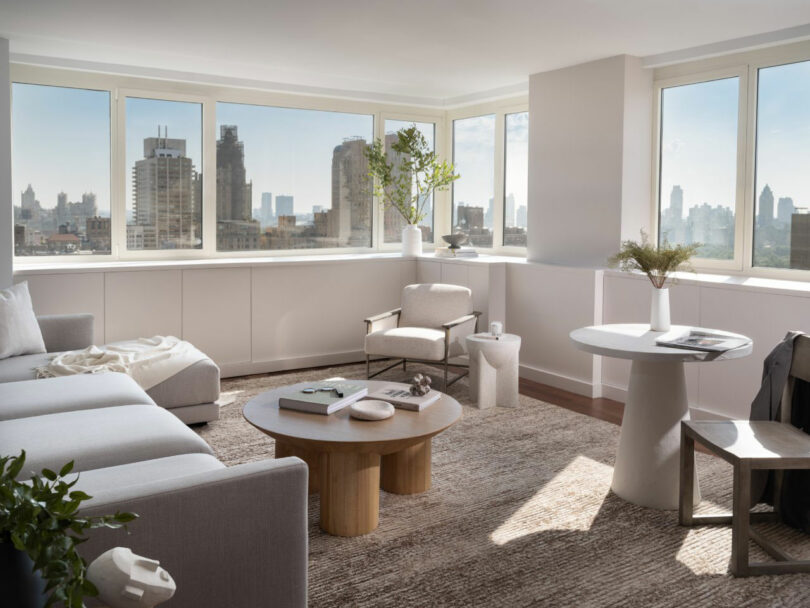 Bright living room with large windows, city view, gray sectional sofa, wooden coffee table, round side table, beige armchair, and decorative plants on a textured rug.