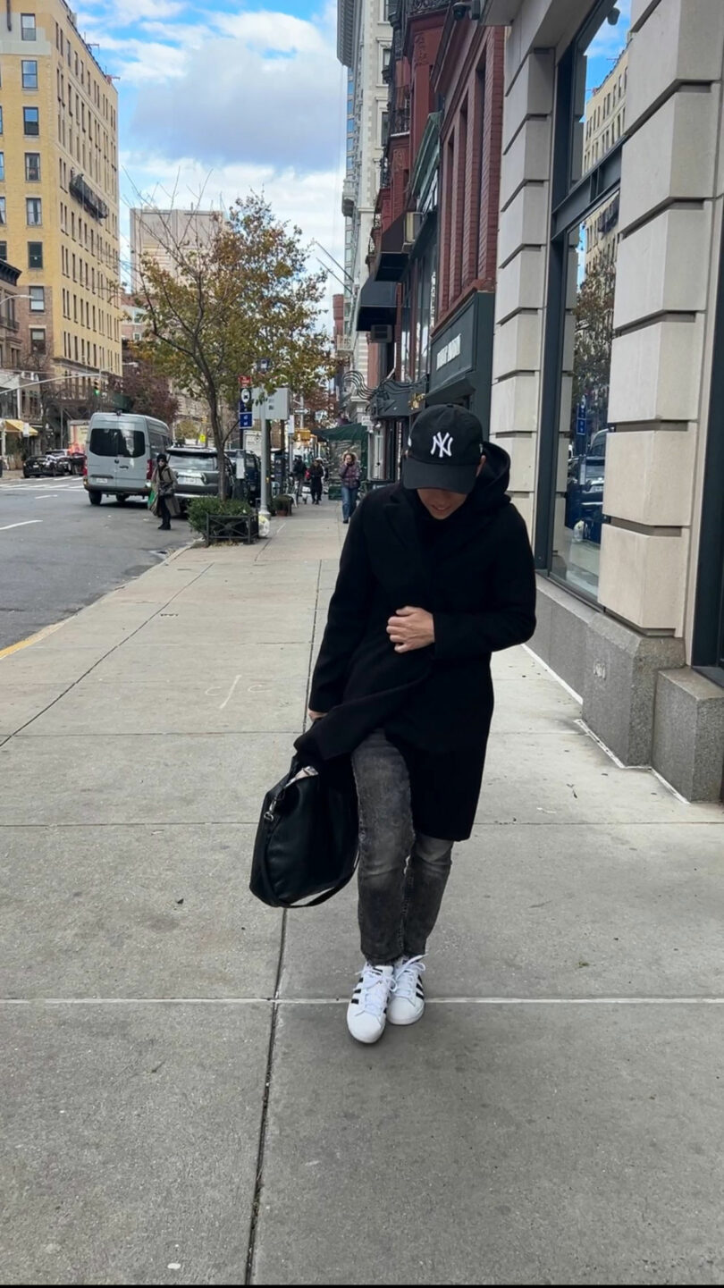 A person in a black coat and cap walks along a city sidewalk, carrying a black bag. Buildings and a parked van are visible in the background.