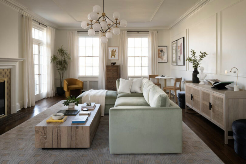 Modern living room with a light green sectional sofa, wooden coffee table, and large windows. A geometric chandelier hangs from the ceiling. Minimalistic decor includes plants and framed art.