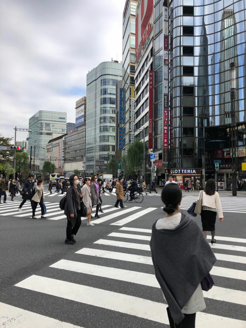 People crossing a bustling city intersection with tall buildings, shops, and signs in the background.