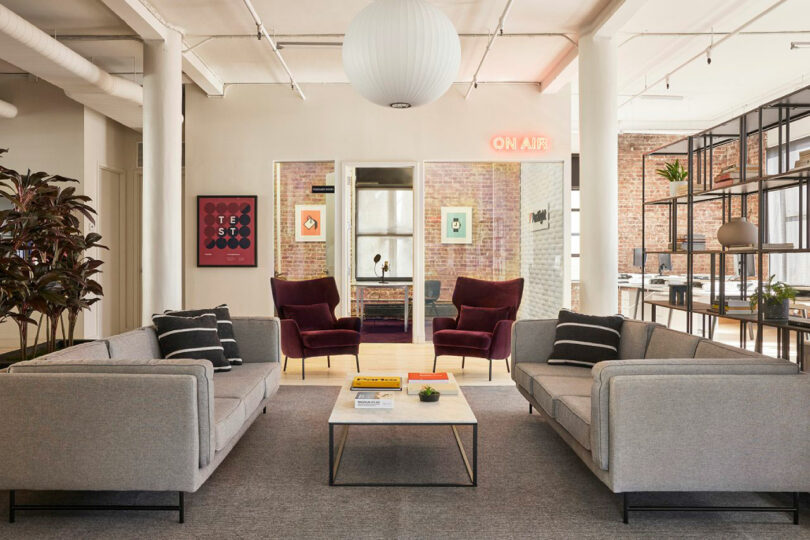 A modern office lounge with two gray sofas and two burgundy armchairs around a low table. A neon 