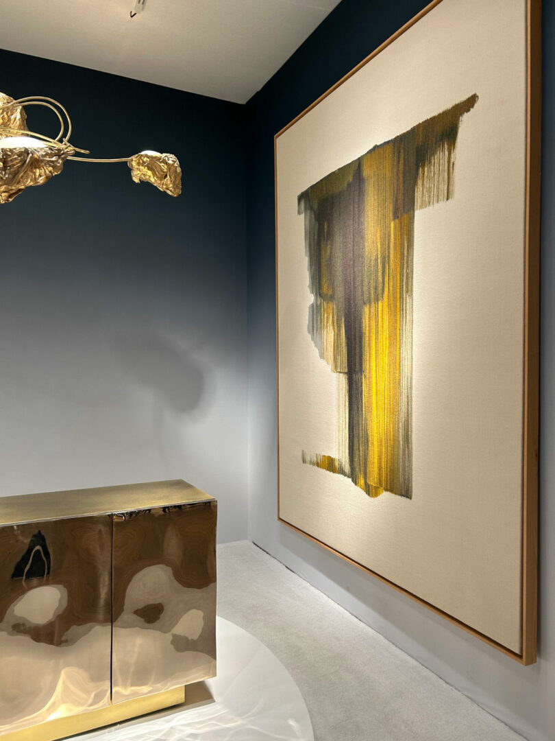 Large abstract painting with vertical yellow and black streaks on a white canvas, hung on a wall. A metallic cabinet and a sculpture with golden leaves are in the foreground.