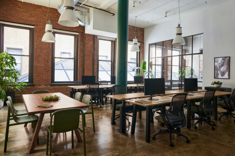 Spacious office with brick walls, large windows, modern desks, chairs, and plants. Gently lit by white ceiling lamps.