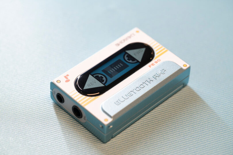 A retro-styled Bluetooth audio player designed to resemble a cassette tape lies on a light blue surface.