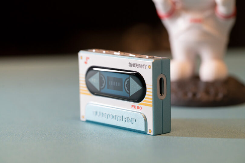 A small, retro-style Bluetooth MP3 player resembling a cassette tape is placed on a blue surface. An astronaut figurine is blurred in the background.