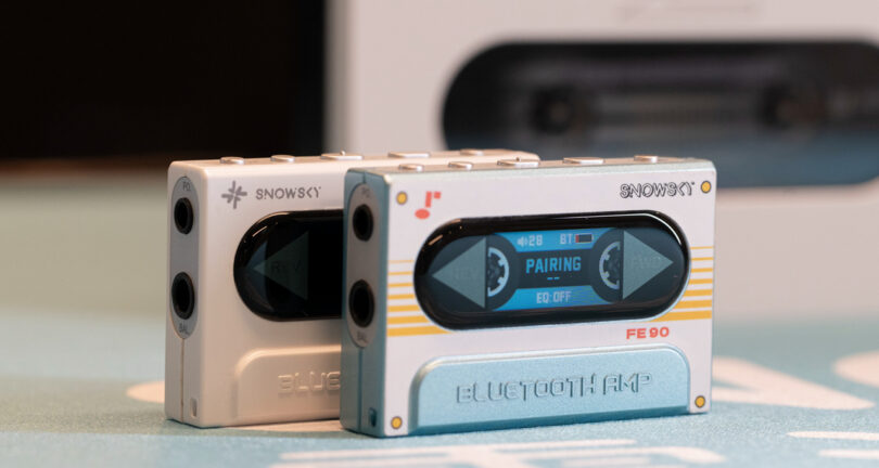 Two compact, retro-style Bluetooth amplifiers with cassette tape design placed on a surface, one displaying 