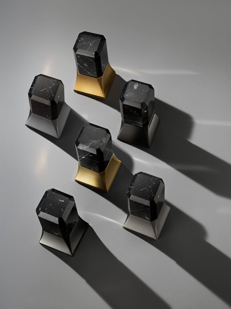 Six geometric black stone objects with gold and metallic bases are arranged on a gray surface, casting long shadows to the right.