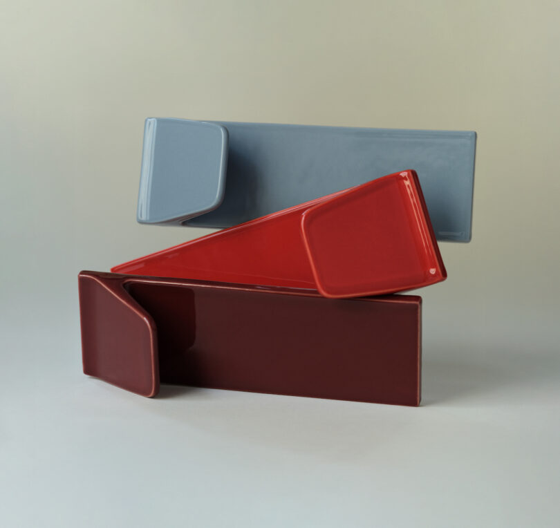 Three rectangular, overlapping ceramic tiles in red, maroon, and gray are arranged in a geometric composition against a neutral background