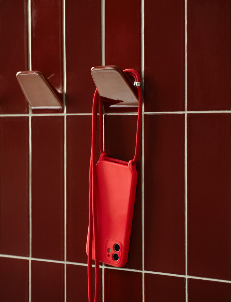 Red smartphone with a matching case and strap hangs on a red tiled wall using two metal hooks