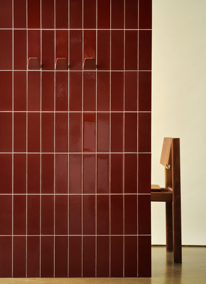 Red-tiled wall with three hooks on the left, adjacent to a wooden chair with a beige backrest, set against a neutral background