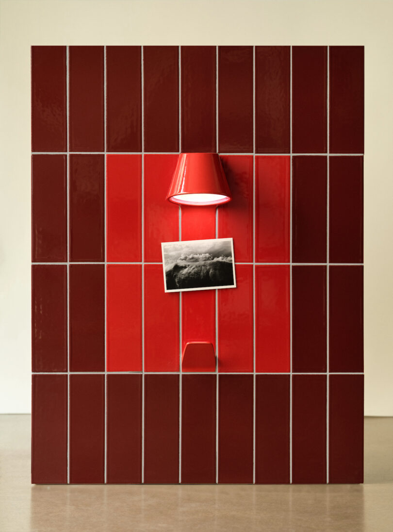 Red tiled wall with a matching red lamp above a black and white photo, centered in the arrangement. Brown floor and plain beige background
