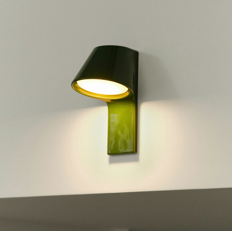 A modern, wall-mounted green lamp with a cylindrical design illuminates a white wall adorned with UU Tiles, casting light in an upward direction.