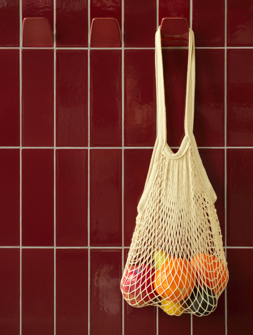 A beige net bag holding oranges and apples hangs against a wall of maroon tiles