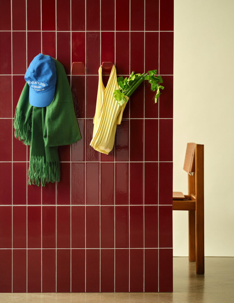 Red tiled wall with hooks holding a blue hat, green scarf, yellow bag, and celery stalks. Wooden chair visible in the background