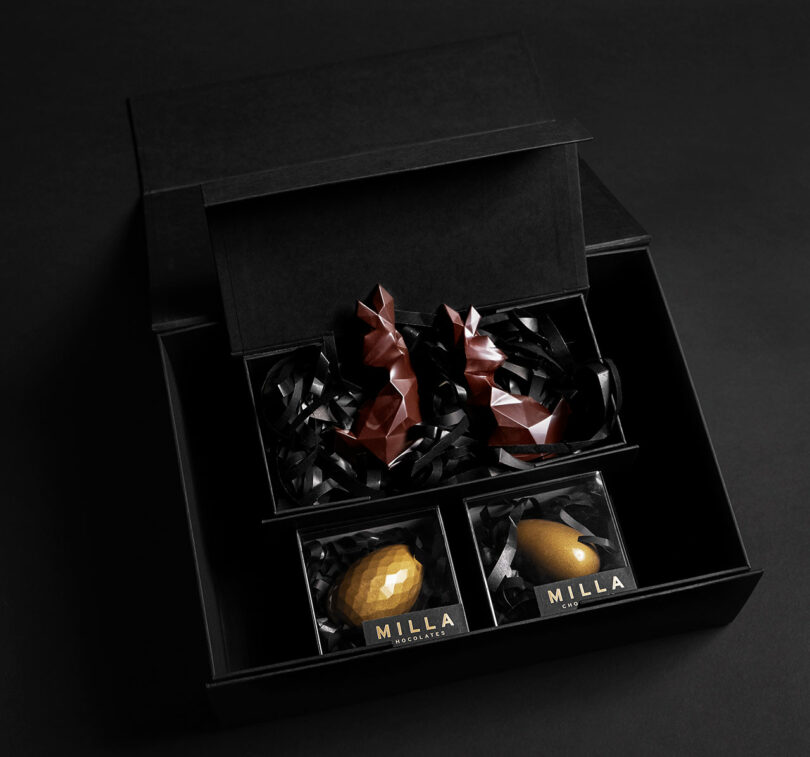 An open black box displaying two geometric brown chocolate bunnies and two smaller golden egg-shaped chocolates in individual boxes labeled