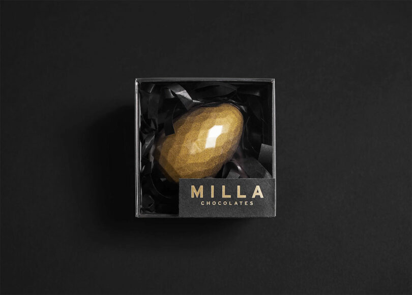 A gold, faceted, gem-shaped chocolate is nestled in black paper inside a black box labeled