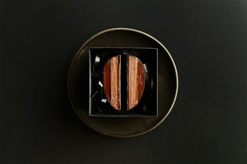 Two glossy, rectangular chocolate pieces are placed side by side in a black square box, on a round dark plate against a dark background.