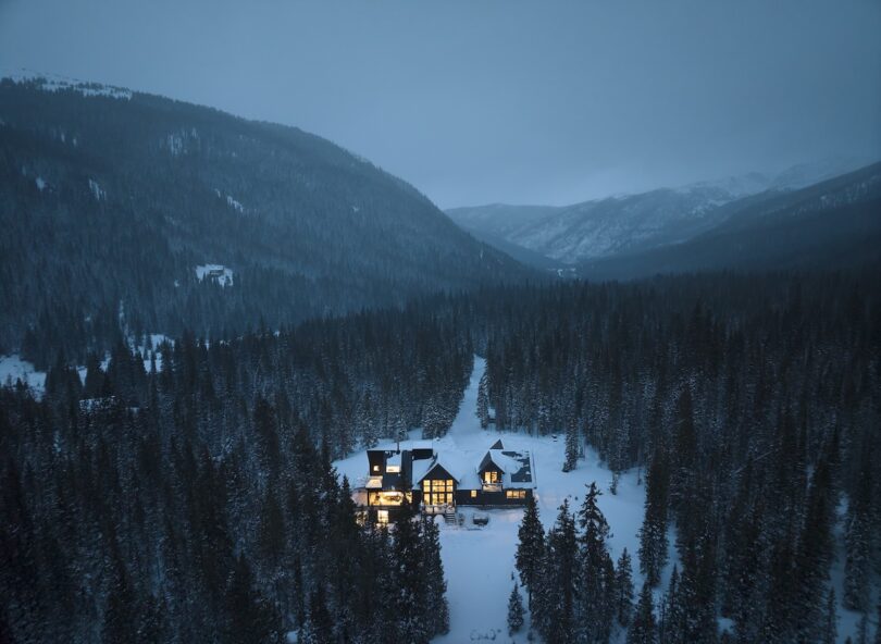 A snow-covered house in a dense forested valley, illuminated warmly at dusk, surrounded by mountains under a dimly lit cloudy sky.