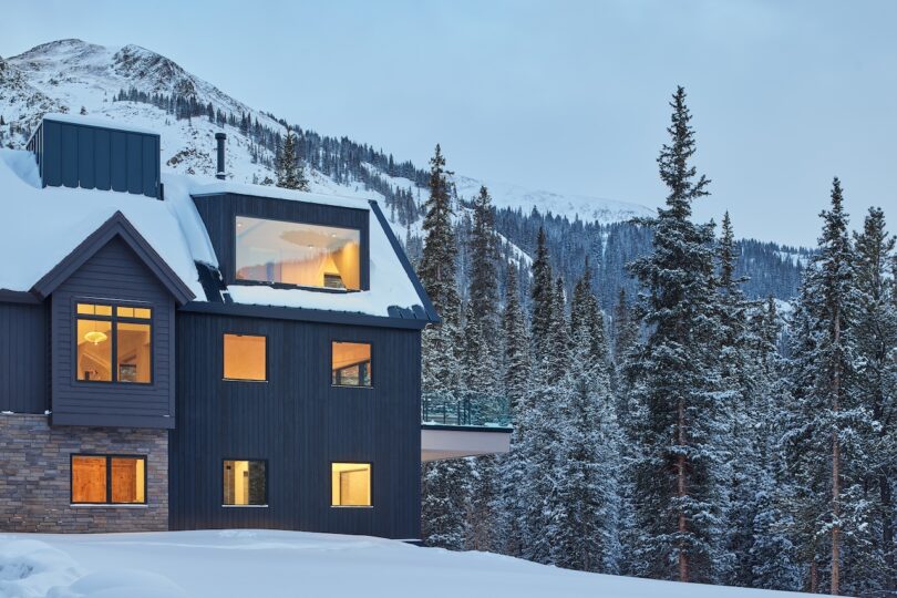 Snow-covered modern house with large windows amid pine trees and mountains in the background, during dusk.