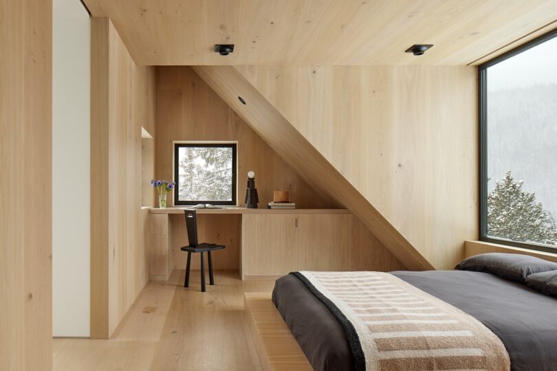 Minimalist bedroom with light wood paneling, large windows, and sloped ceiling. A bed with dark bedding, a chair, and a small desk with decor are present. Snowy trees visible outside.