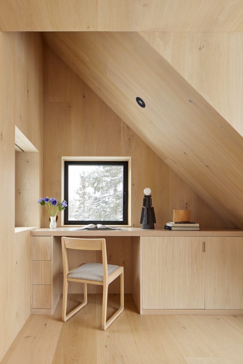Minimalist wooden workspace under a sloped ceiling, featuring a desk with a chair, books, a small sculpture, a window view of trees, and a vase with purple flowers.