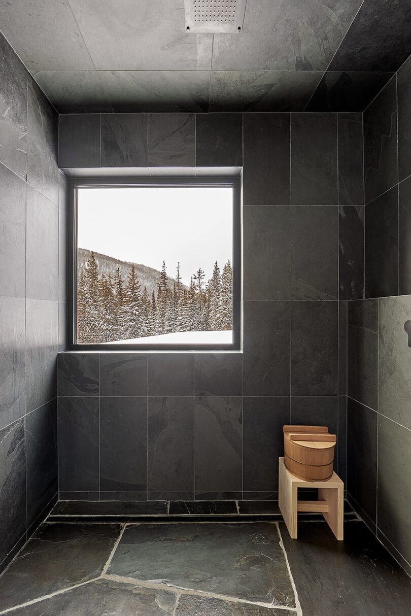 Slate-tiled shower room with a wooden stool and bucket, featuring a large window offering a snowy forest view.