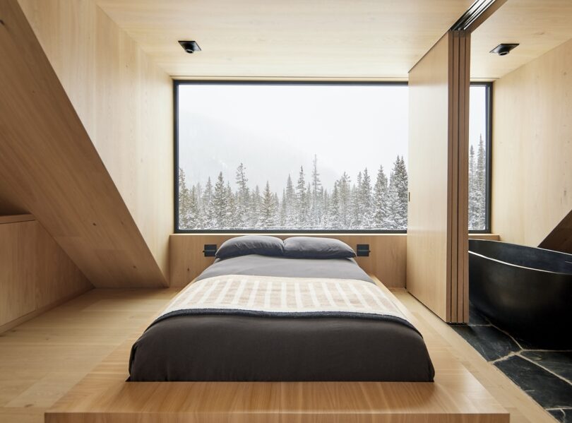Minimalist bedroom with a wooden interior, featuring a bed facing a large window. Snowy forest landscape is visible outside.
