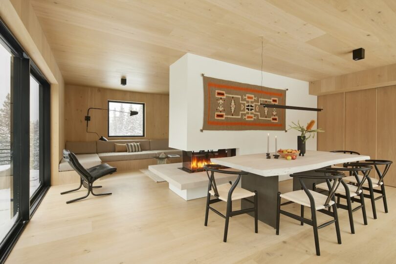 Modern living and dining area with light wood floors, a central fireplace, a cushioned seating nook, black chair, and a dining table with black chairs. Large windows and a woven wall decoration.
