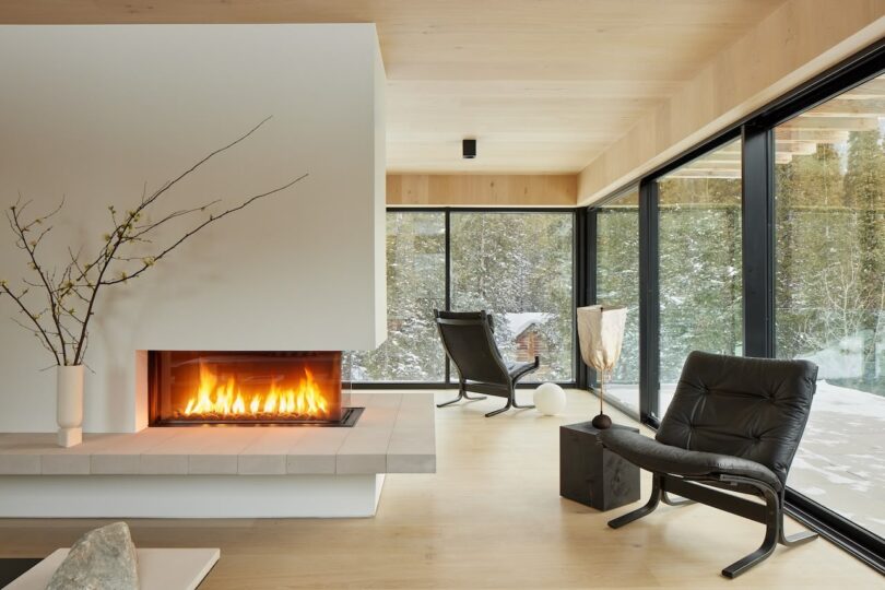 Modern living room with large windows, a lit fireplace, minimalist décor, and two black chairs. Snowy forest view outside.
