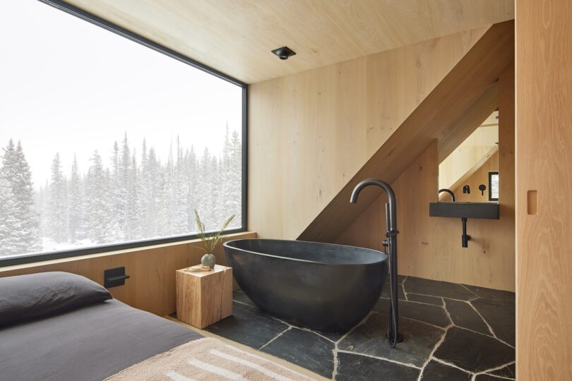Modern bedroom-bathroom with a black freestanding tub, wooden walls, large window with snowy forest view, small wooden side table, and a black faucet by a geometric mirror and sink.