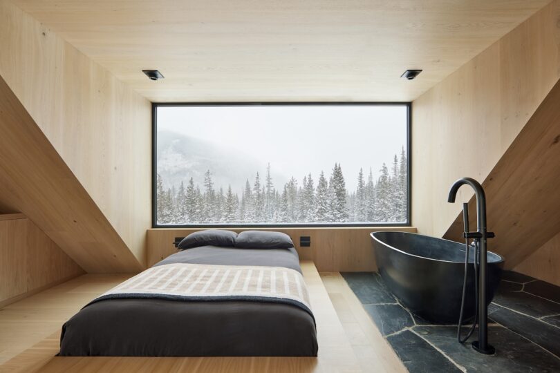 Minimalist bedroom with a large window overlooking snowy trees. Features a single bed, freestanding black bathtub, and wood-paneled walls and floor.