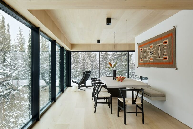 Minimalist dining room with large windows overlooking snowy trees. A long table, chairs, a bench, a wall tapestry, and a black reclining chair adorn the space.