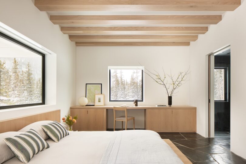 Modern bedroom with a large bed, wooden beams, and a desk under two windows. A vase with branches and framed pictures decorate the room, overlooking snow-covered trees outside.