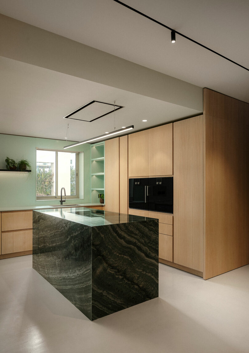 Modern kitchen with light wood cabinets, black integrated appliances, and a large green marble island. The room is illuminated by track lighting and has a window above the sink.