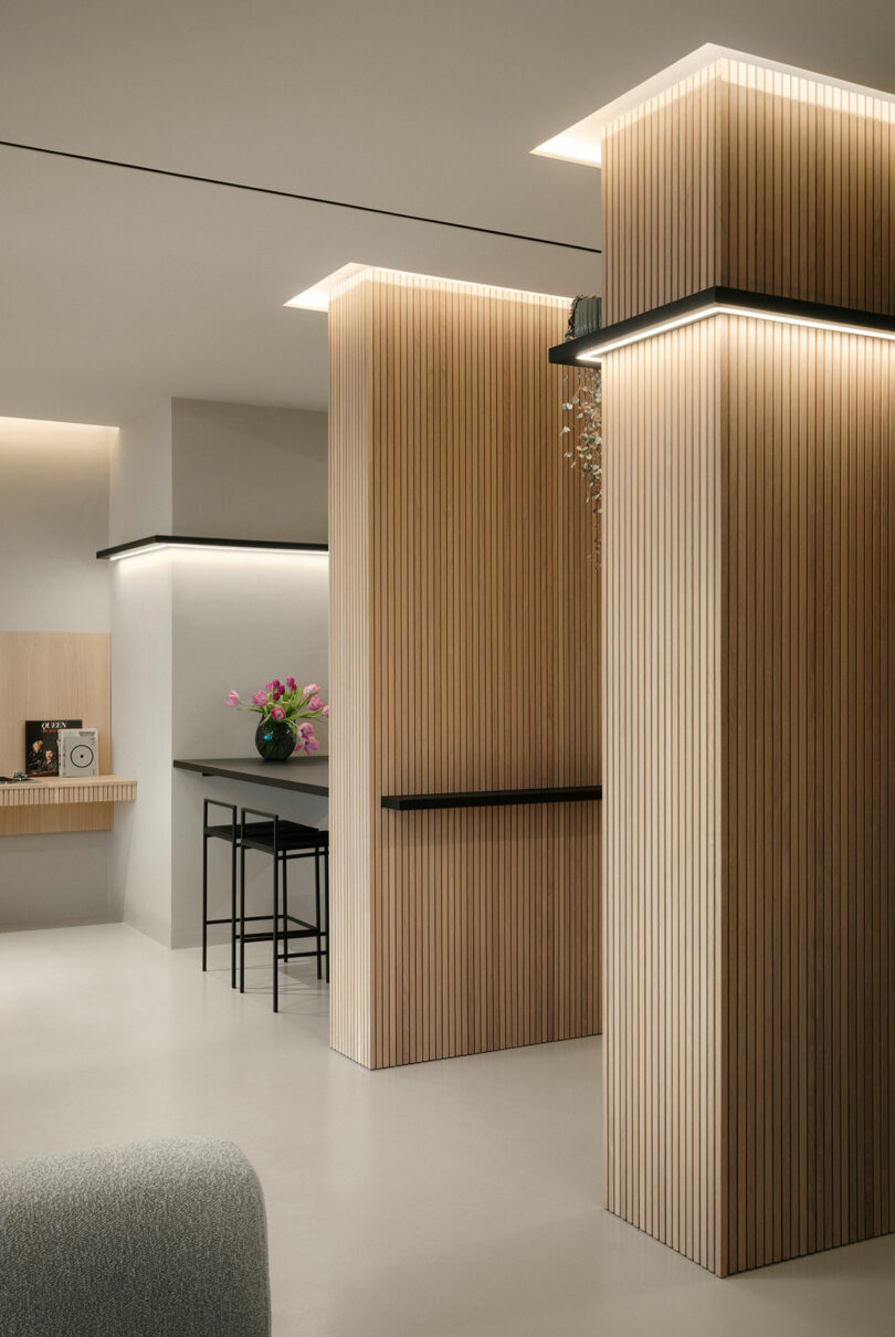 Modern interior with vertical wooden panels, soft lighting, a small desk with a chair, and a vase of pink flowers. Minimalist aesthetic with a neutral color palette.