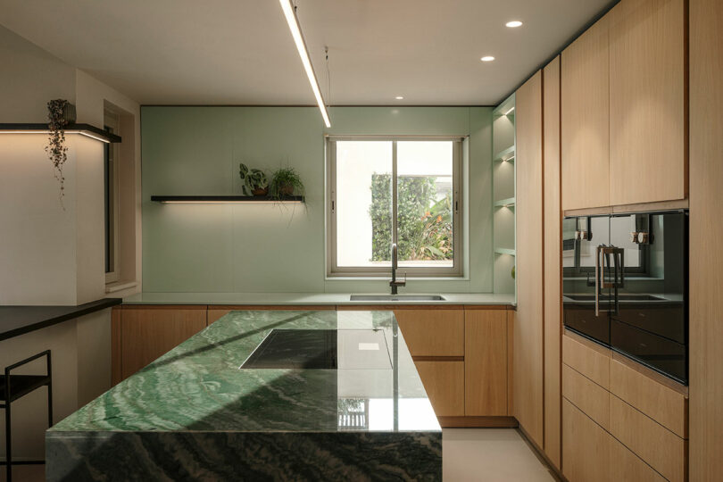 Modern kitchen with light wood cabinetry, green marble island, built-in oven, induction cooktop, and wall shelves. A large window provides natural light.