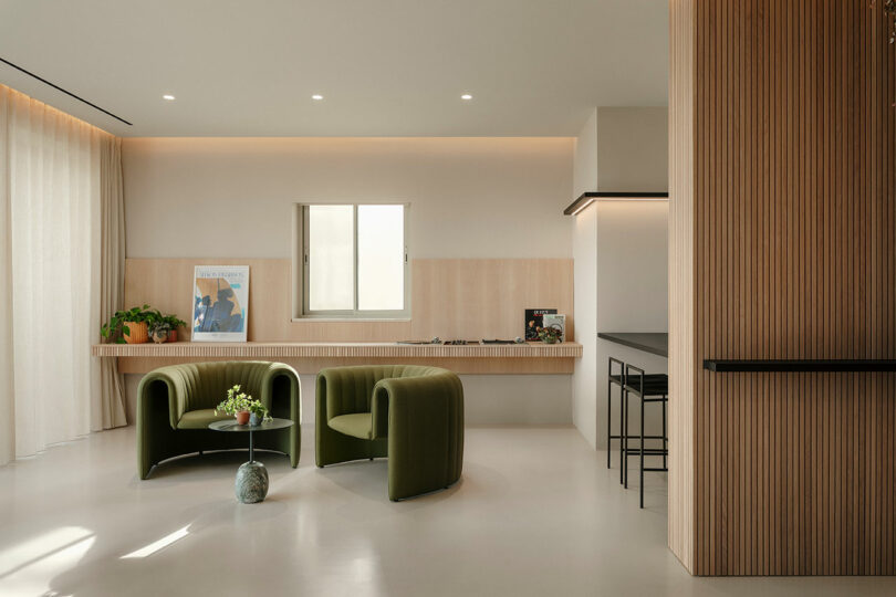 Minimalist living room with two green chairs, a small round table with a plant, and a wooden bench under a window. A magazine and plants decorate the space. A kitchen area is visible on the right.