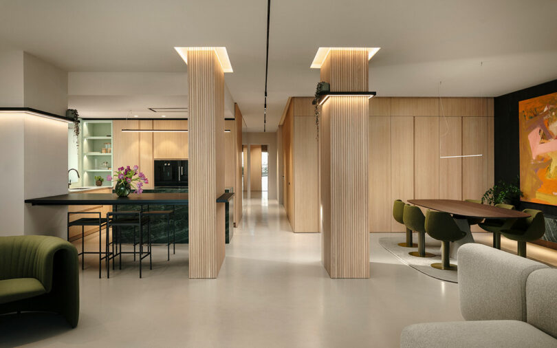 Modern open-plan interior with wood-paneled columns, a kitchen island, dining table, green chairs, and abstract wall art.