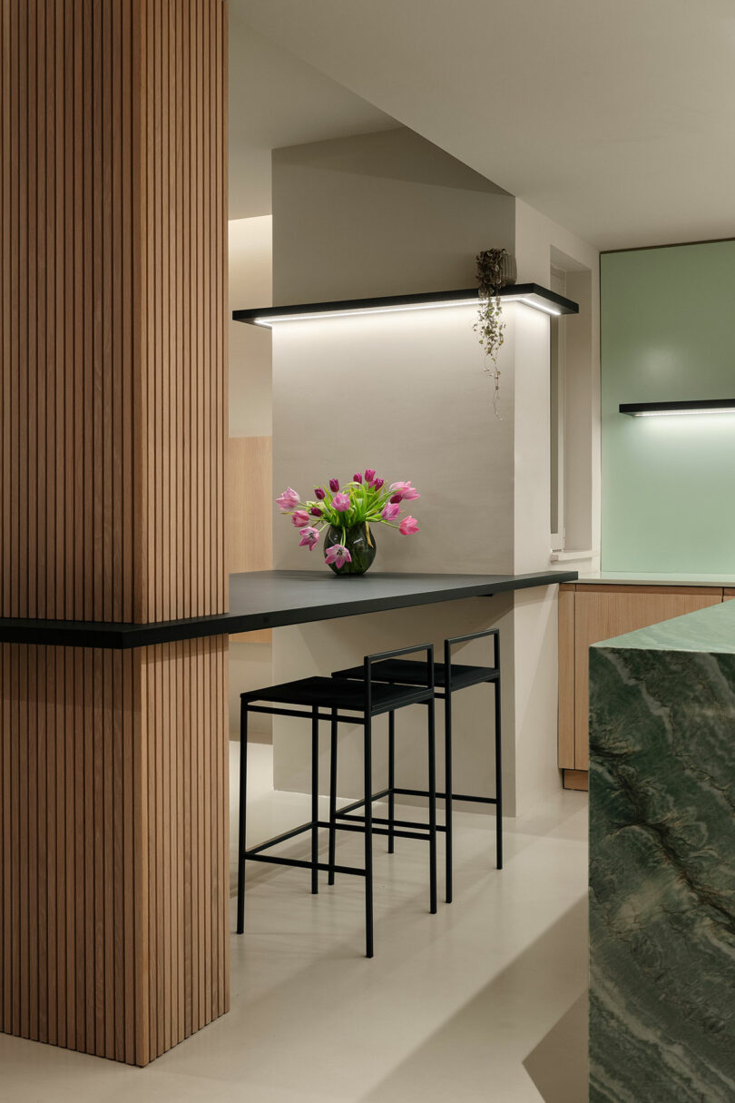 Modern kitchen interior with a black countertop and two bar stools, featuring a vase of pink tulips. Wooden panels and a green marble surface add contrast to the minimalist design.