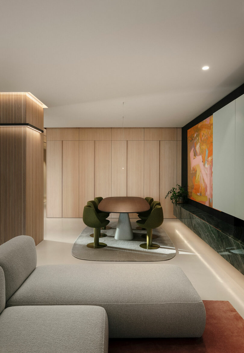 Modern minimalist living room with a gray sectional sofa, green upholstered dining chairs around a brown table, large abstract wall art, and wood paneling.