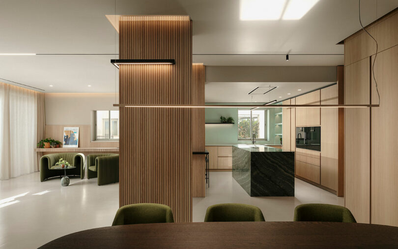 Modern kitchen and dining area with wood paneling, green chairs, marble island, and large windows, creating an open and bright space.