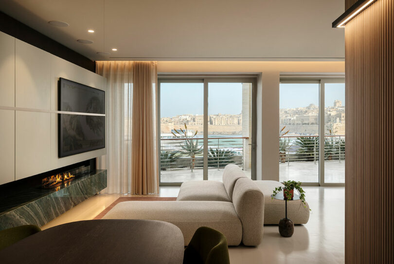 Modern living room with a sleek sofa, wall-mounted TV above a fireplace, and large windows overlooking a waterfront view. A small plant sits beside the sofa.