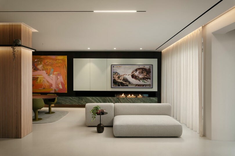 Modern living room with minimalist furniture, abstract art, and a linear fireplace. Features a soft gray sofa, wood accents, green table chairs, and large white curtains.