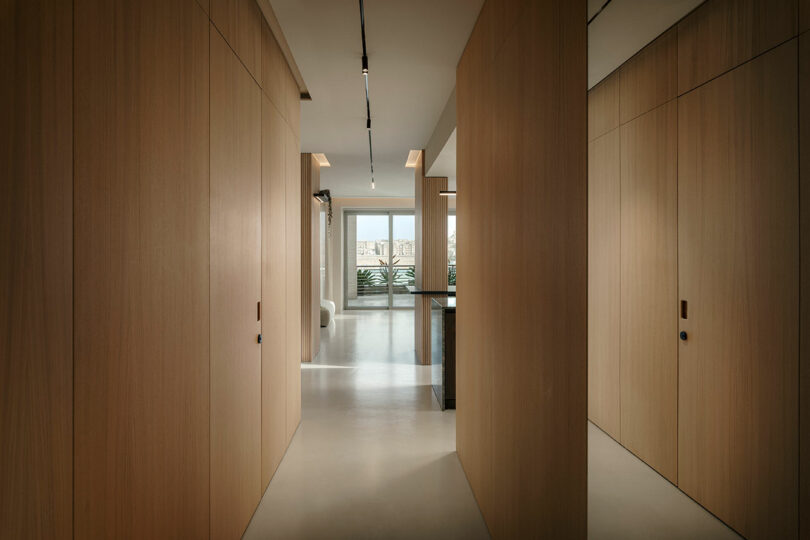 Minimalist hallway with wooden panel walls, recessed lighting, and a distant window view of a balcony with greenery.