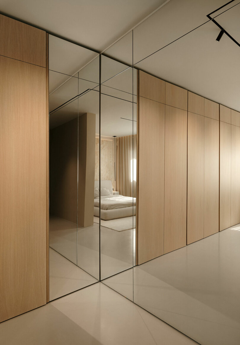 A minimalist bedroom with light wood paneling and mirrored doors, leading to a bed with white linens.