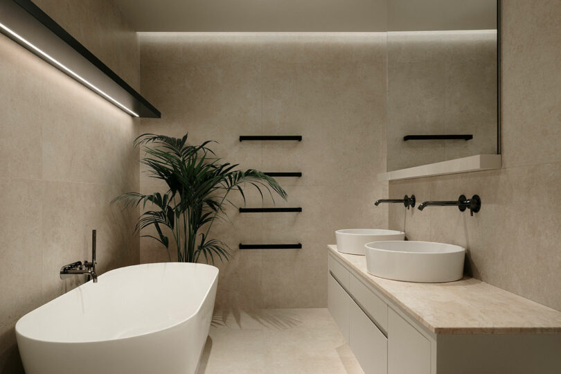 Minimalist bathroom with a freestanding bathtub, double sink vanity, and a large mirror. Beige tile walls and floor, with a potted plant and modern black fixtures.